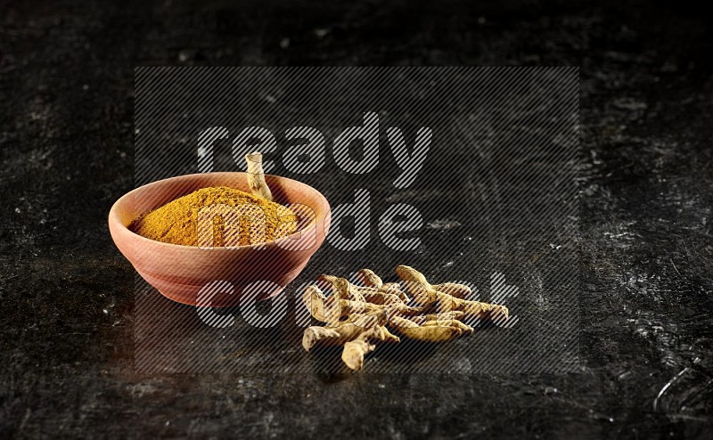 A wooden bowl full of turmeric powder with dried turmeric whole fingers on textured black flooring