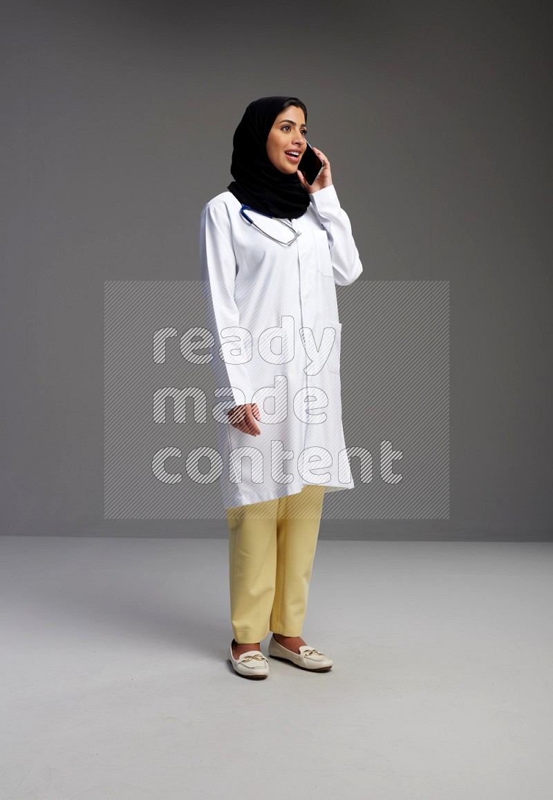 Saudi woman wearing lab coat with stethoscope standing talking on phone on Gray background