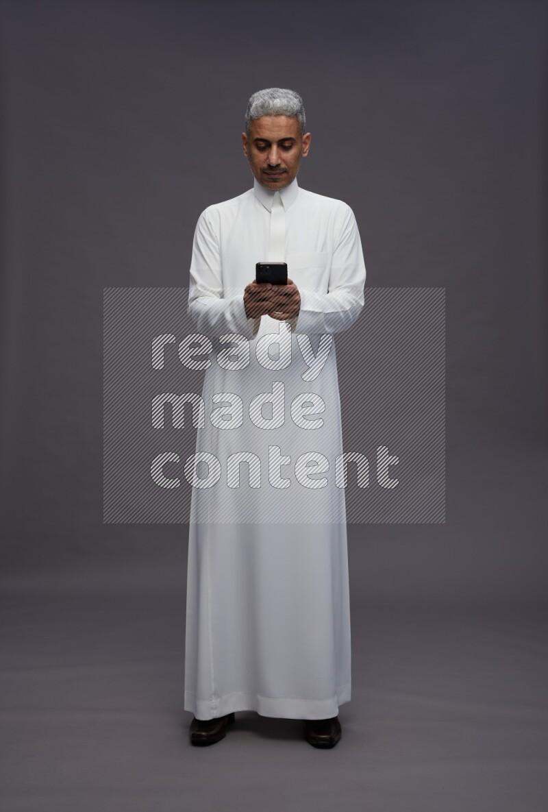 Saudi man wearing thob standing texting on phone on gray background