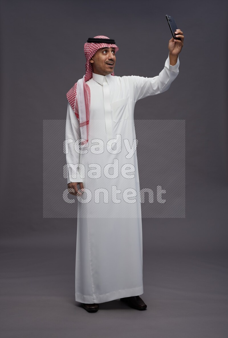 Saudi man wearing thob and shomag standing taking selfie on gray background
