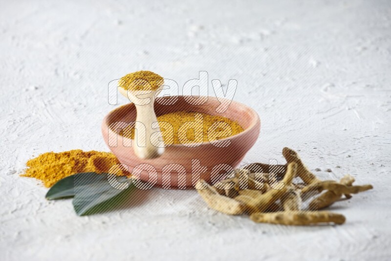 A wooden bowl and wooden spoon full of turmeric powder with dried turmeric fingers beside it on textured white flooring