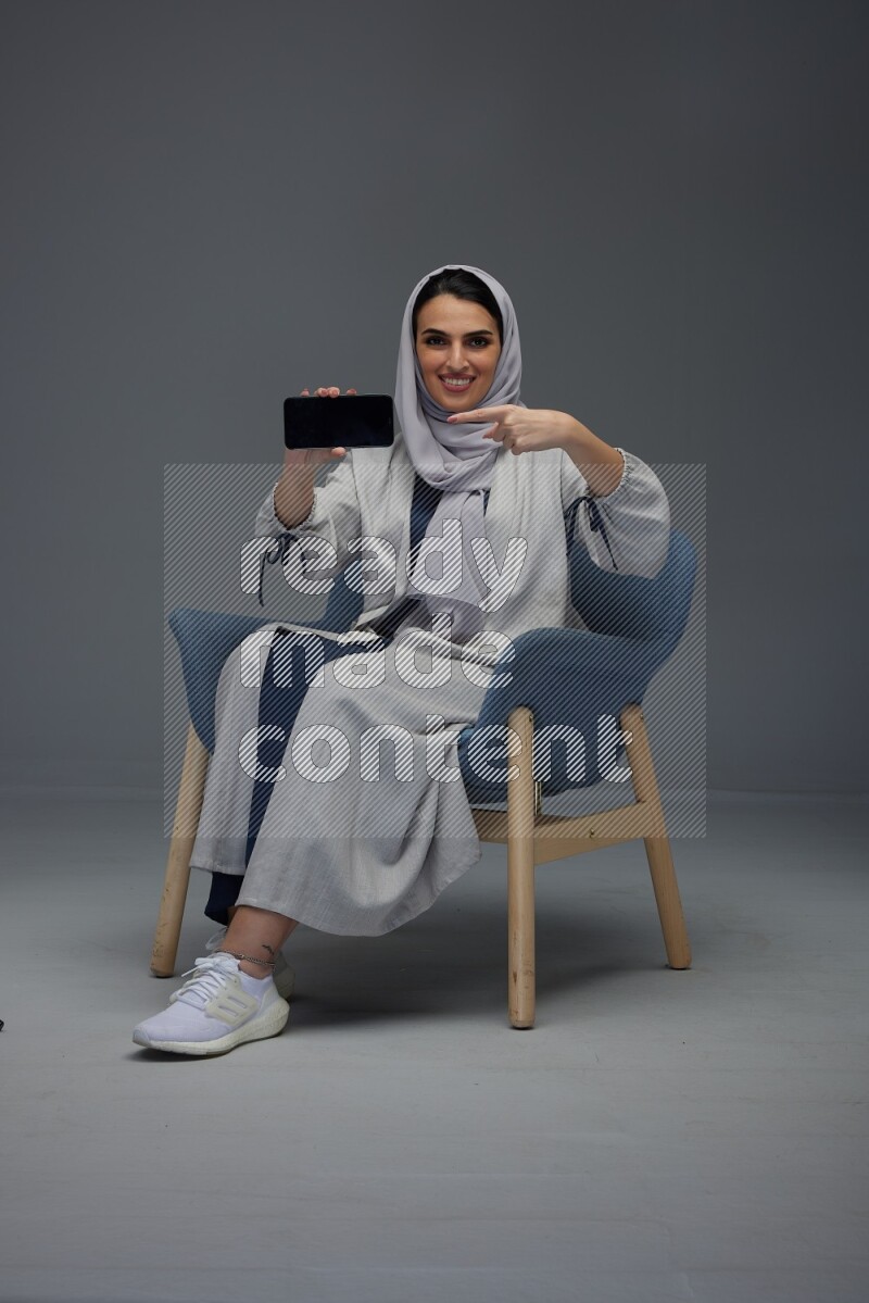 A Saudi woman wearing a light gray Abaya and head scarf sitting on a dark grey chair and showing the phone's screen eye level on a grey background
