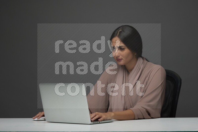 A Saudi woman Sitting on her desk on a Gray Background wearing Brown Abaya
