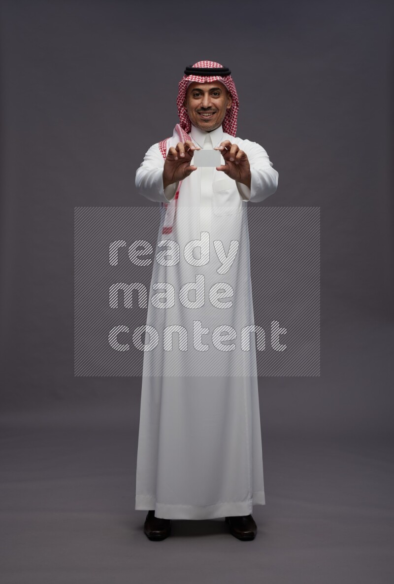 Saudi man wearing thob and shomag standing holding ATM card on gray background