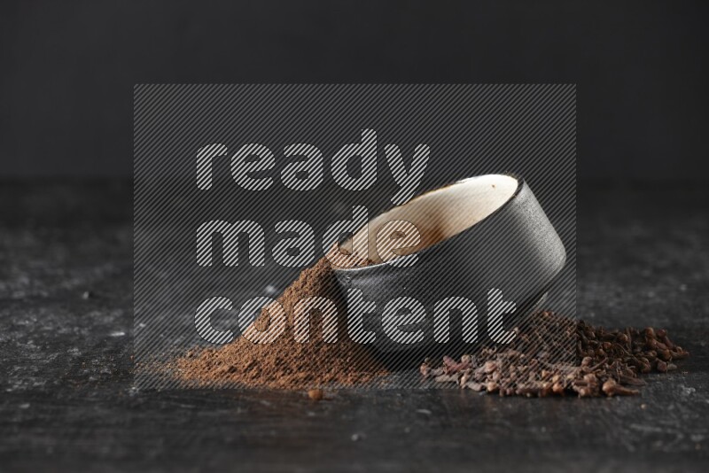 وعاء فخاري أسود ممتلئ ببودرة القرنفل والبودرة تناثرت منه مع الحبوب على خلفية سوداء