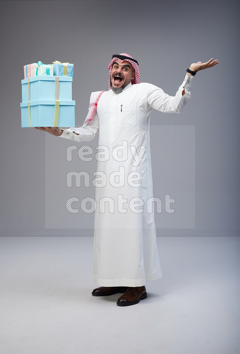 Saudi man Wearing Thob and red Shomag standing holding gift box on Gray background