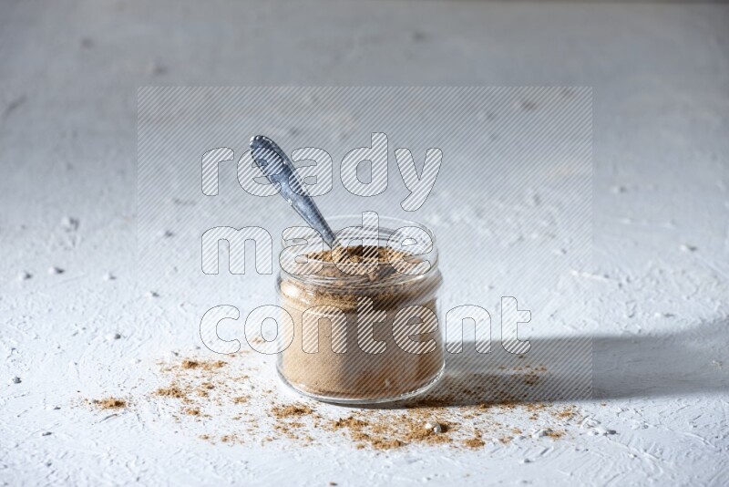 A glass jar and a metal spoon full of allspice powder on a textured white flooring