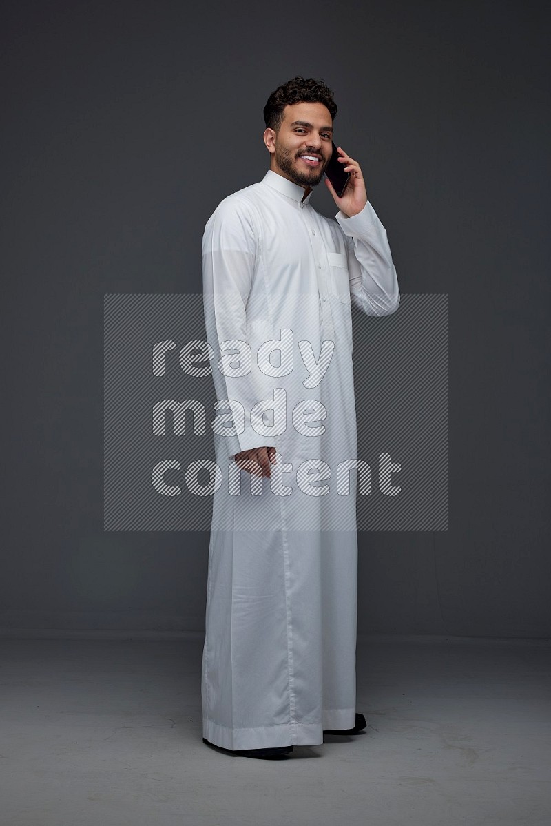 A Saudi man wearing Thobe and talking in the phone while standing and making different hand gestures eye level on a gray background