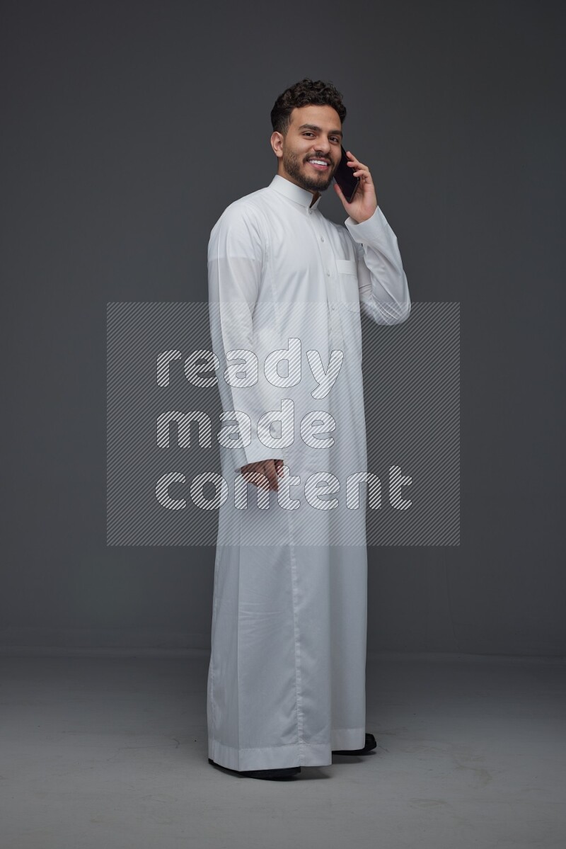 A Saudi man wearing Thobe and talking in the phone while standing and making different hand gestures eye level on a gray background