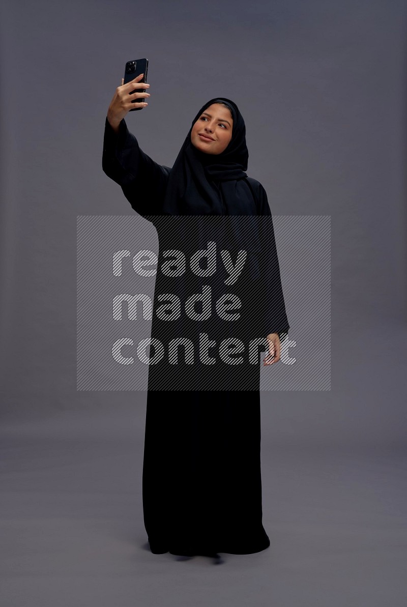 Saudi woman wearing Abaya standing taking selfie on gray background