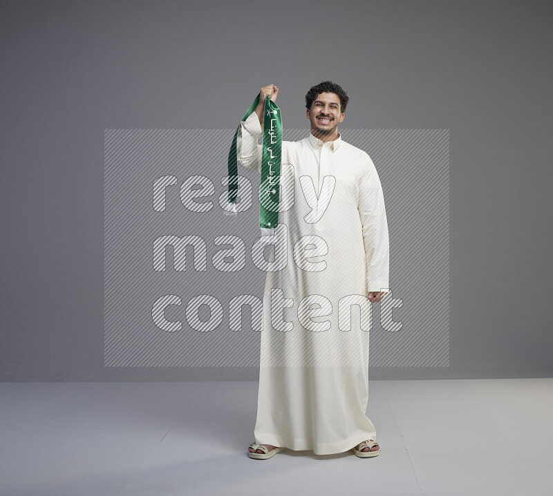 A Saudi man standing wearing thob holding Saudi flag scarf on gray background