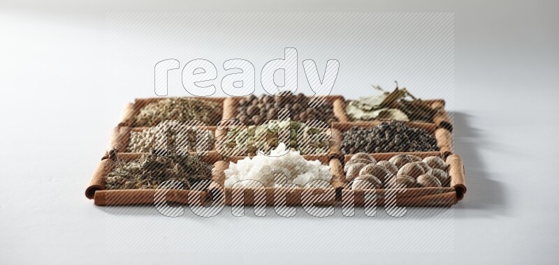 9 مربعات من أعواد القرفة ممتلئة بالهيل في المنتصف محاطة بجوزة الطيب والملح والريحان المجفف والفلفل الأبيض والكمون وبهار الفلفل الحلو وأوراق الغار والفلفل الأسود على أرضية بيضاء