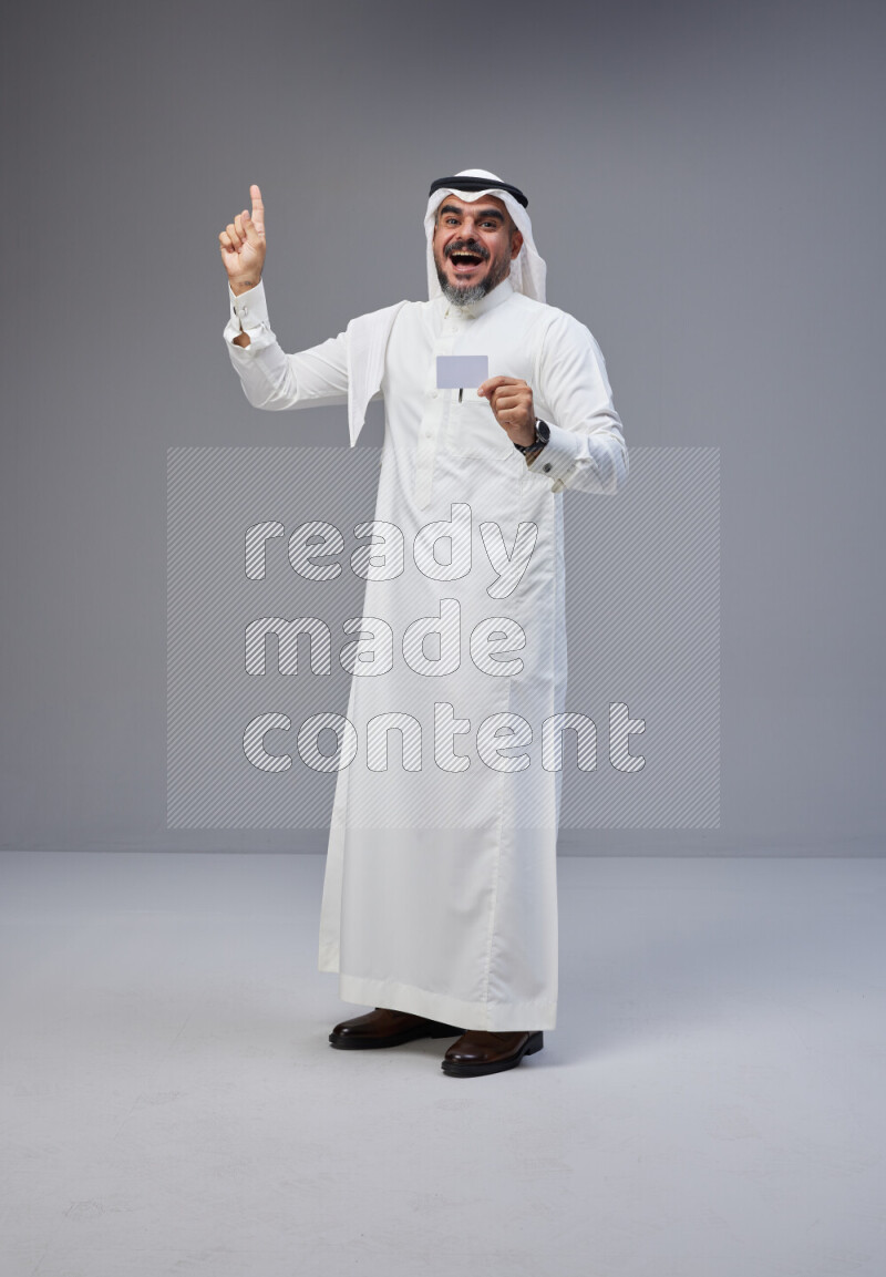 Saudi man Wearing Thob and white Shomag standing holding ATM card on Gray background