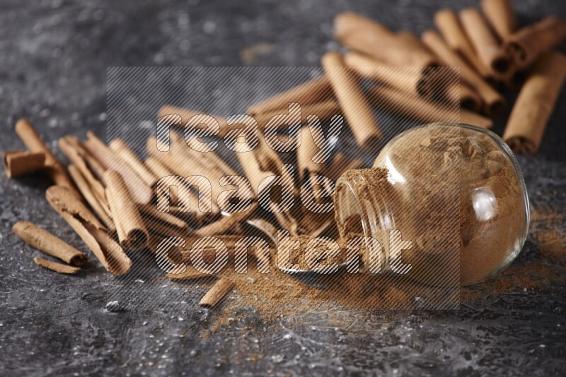 Herbal glass jar full cinnamon powder flipped and a metal spoon full of powder surrounded by cinnamon sticks on textured black background in different angles