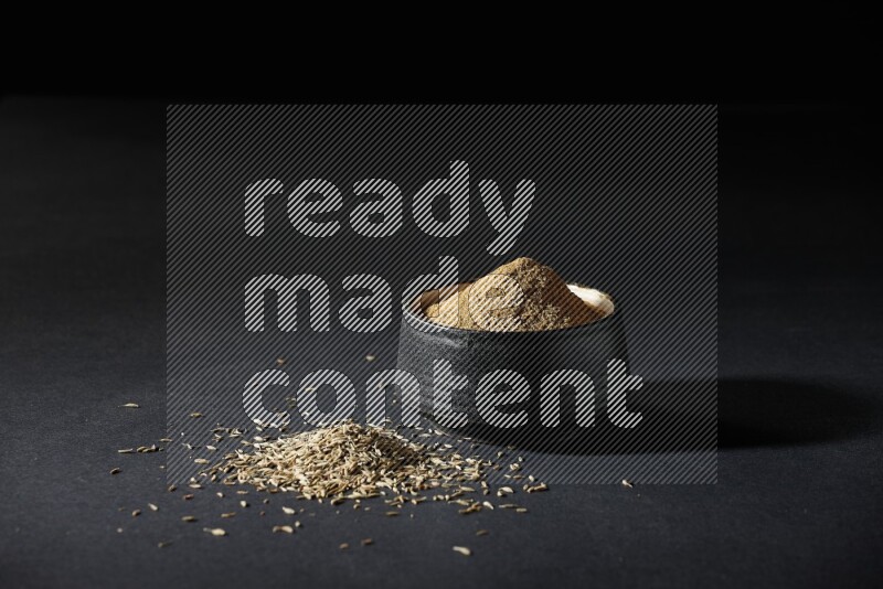 وعاء فخاري أسود ممتلئ ببودرة الكمون وحبوب الكمون متناثرة على خلفية سوداء