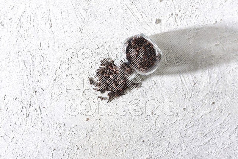 A flipped glass spice jar full of cloves and cloves came out of it on textured white flooring