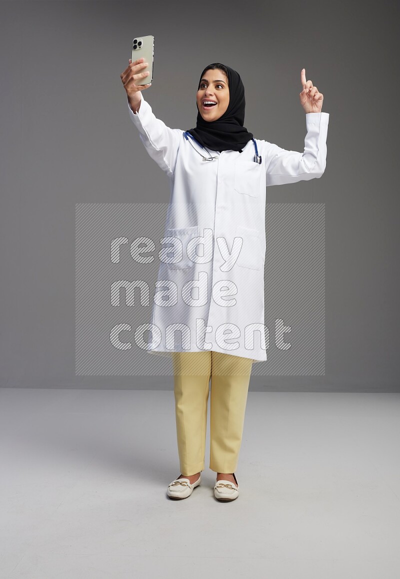 Saudi woman wearing lab coat with stethoscope standing taking selfie on Gray background