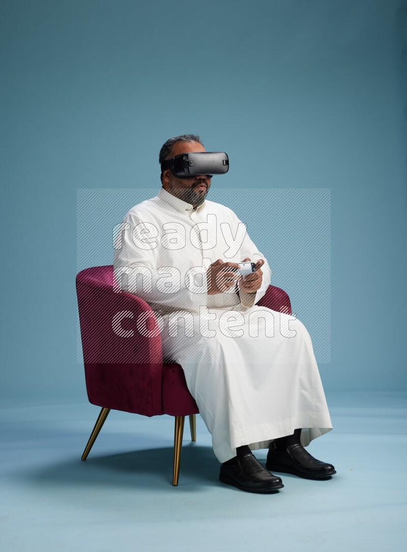 Saudi Man without shimag sitting on chair  gaming with VR and joystick on blue background