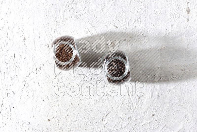 2 glass jars full of cloves powder and cloves grains on a textured white flooring