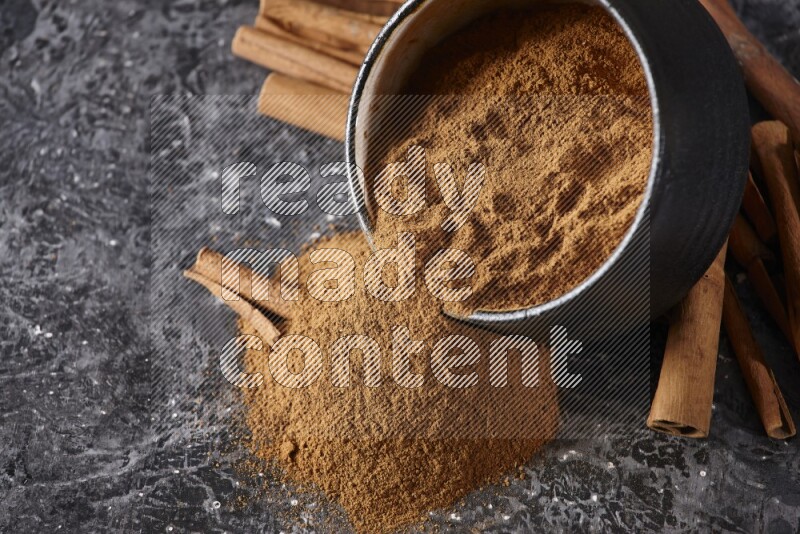 Black pottery bowl over filled with cinnamon powder and cinnamon sticks around the bowl on a textured black background
