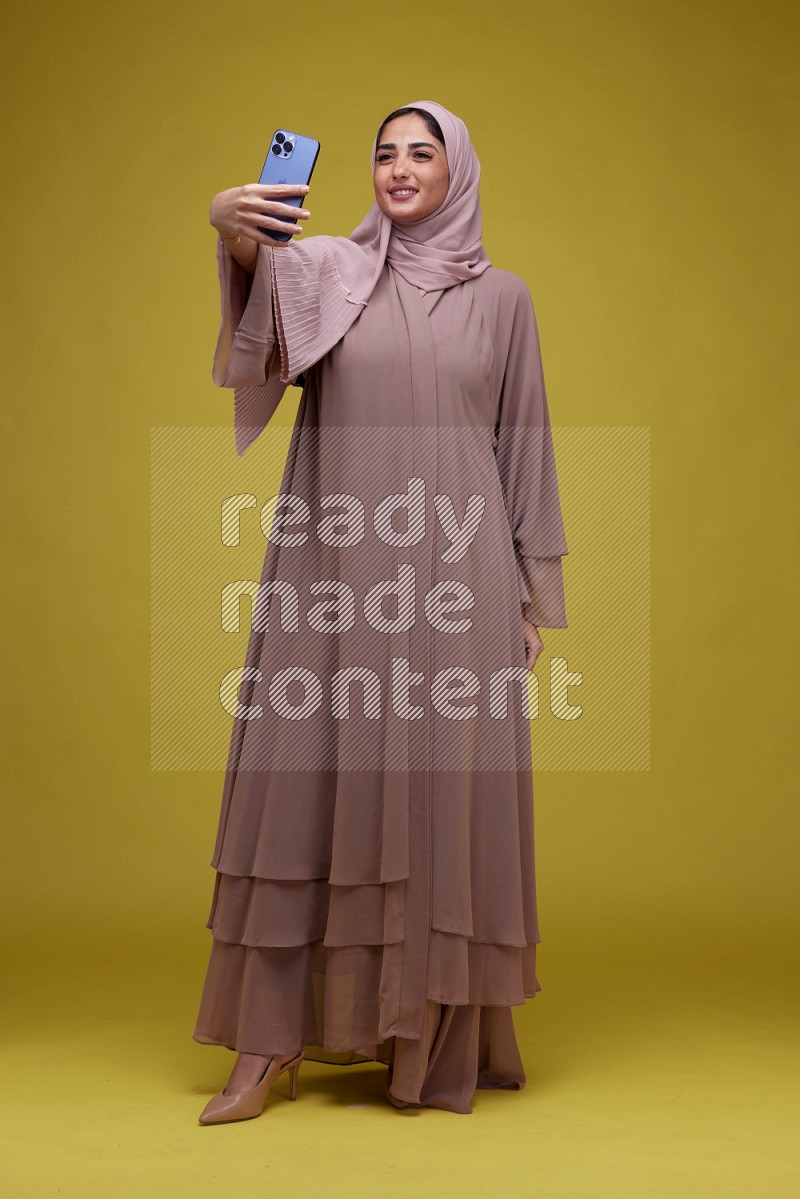 A woman taking a Selfie on a Yellow Background wearing Brown Abaya with Hijab