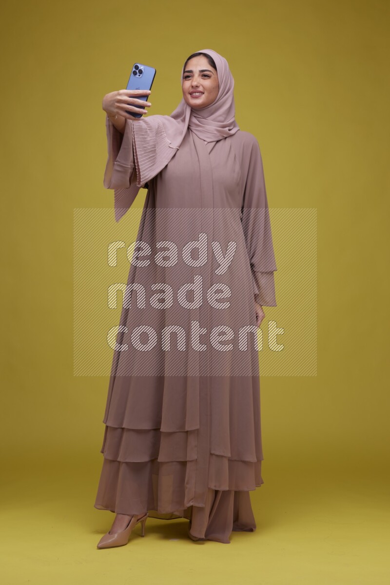 A woman taking a Selfie on a Yellow Background wearing Brown Abaya with Hijab