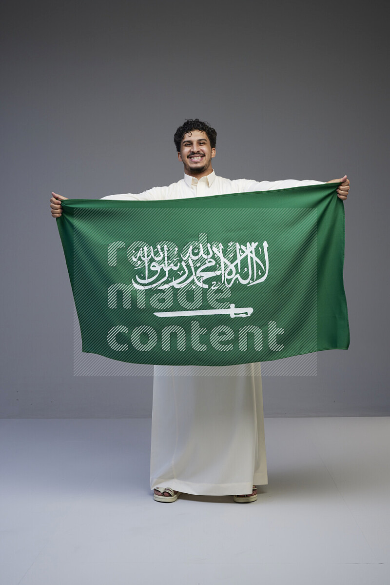 A Saudi man standing wearing thob holding big Saudi flag on gray background