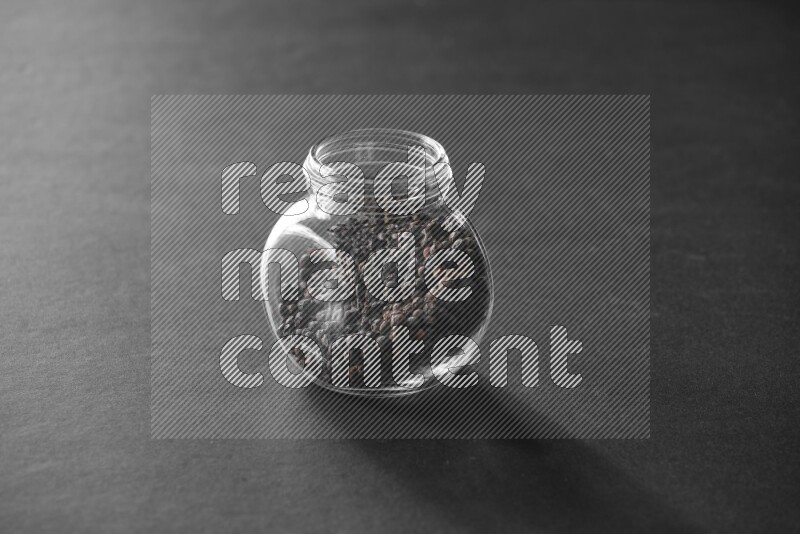 A glass spice jar full of black peppers on black flooring