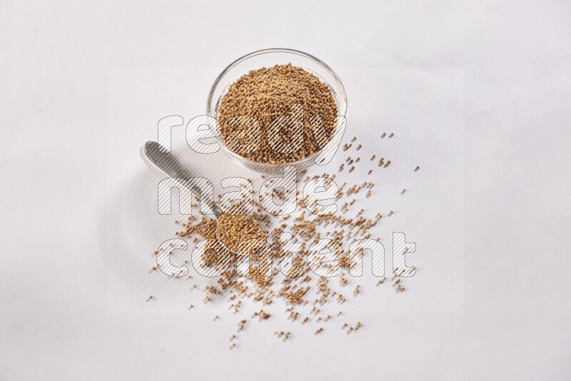 A glass bowl and metal spoon full of mustard seeds on a white flooring