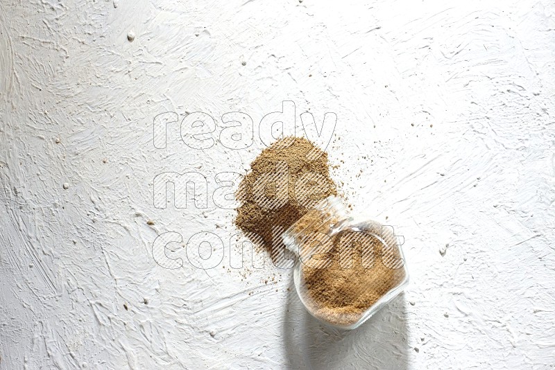 A flipped glass spice jar full of cumin powder and powder spilled out on textured white flooring