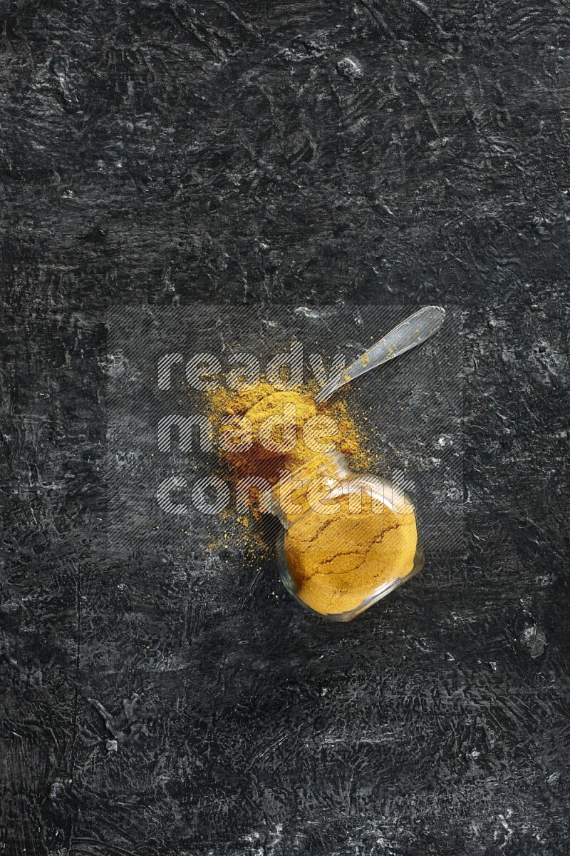 A flipped glass spice jar and a metal spoon full of turmeric powder and powder spilled out of it on textured black flooring