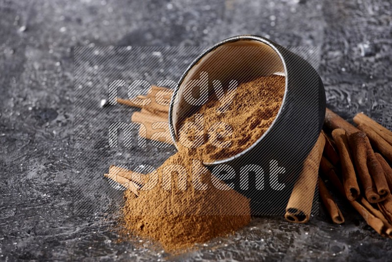 Black pottery bowl over filled with cinnamon powder and cinnamon sticks around the bowl on a textured black background