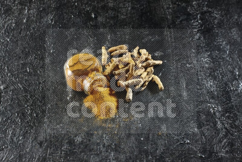 وعاء زجاجي للتوابل ممتلئ ببودرة الكركم مقلوب والبودرة تناثرت منه مع أصابع الكركم المجففة على خلفية سوداء