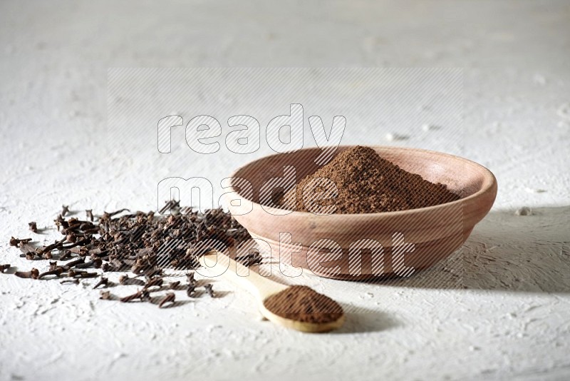 A wooden bowl and wooden spoon full of cloves powder with cloves spread on textured white flooring