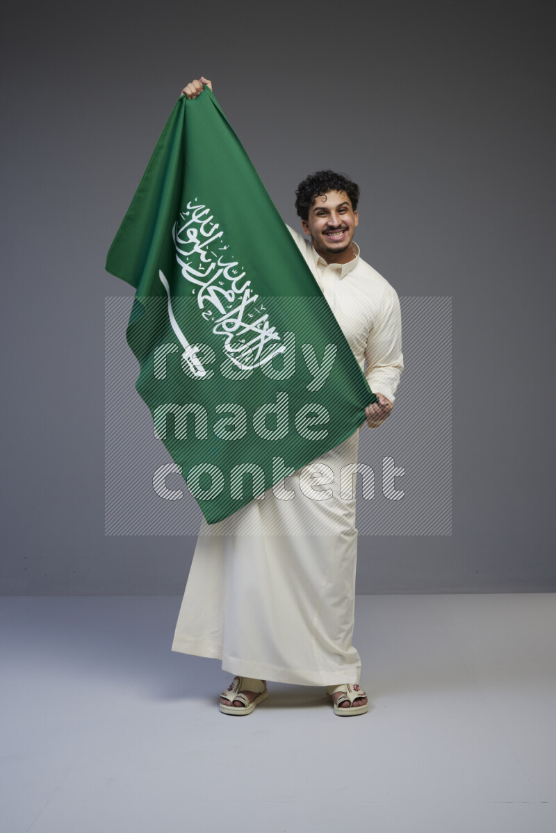 A saudi man standing wearing thob holding big saudi flag on gray background