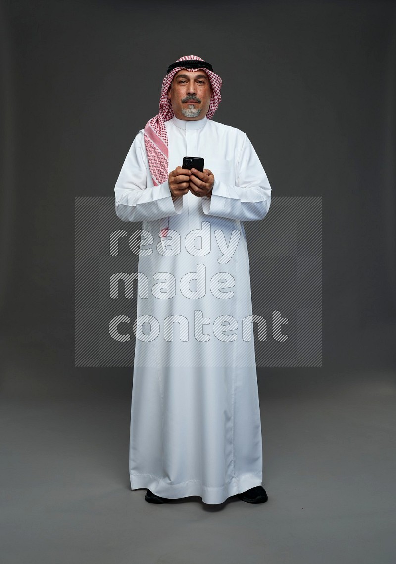 Saudi man with shomag Standing texting on phone on gray background