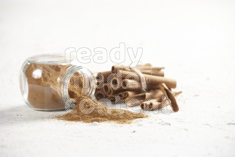 Flipped herbs glass jar full of cinnamon powder and cinnamon sticks in the back on a textured white background