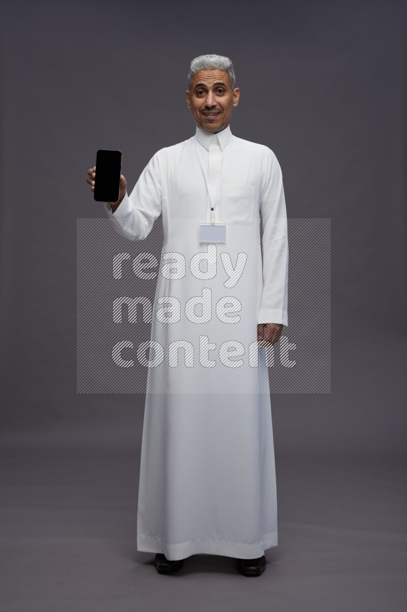 Saudi man wearing thob with neck strap employee badge standing showing phone to camera on gray background