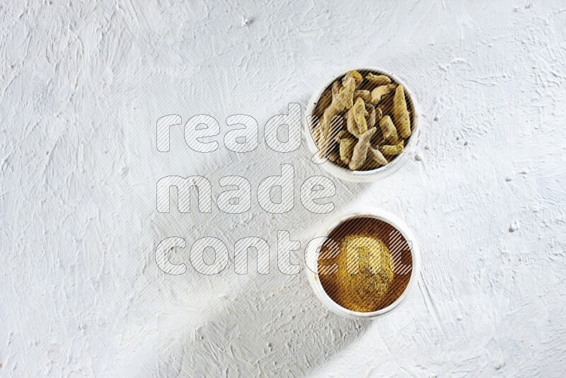 2 beige pottery bowl full of turmeric powder and dried turmeric whole fingers on textured white flooring