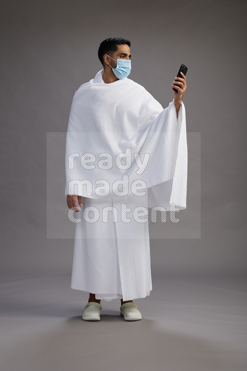 A man wearing Ehram with face mask Standing taking selfie on gray background