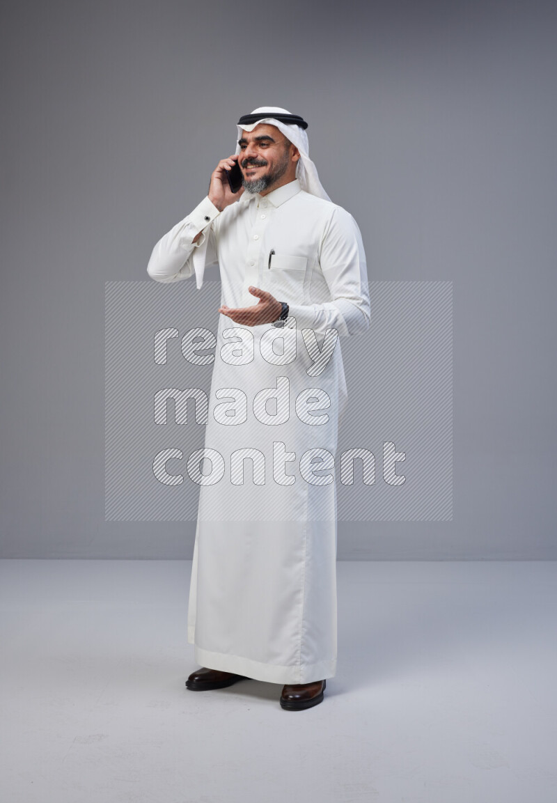 Saudi man Wearing Thob and white Shomag standing talking on phone on Gray background