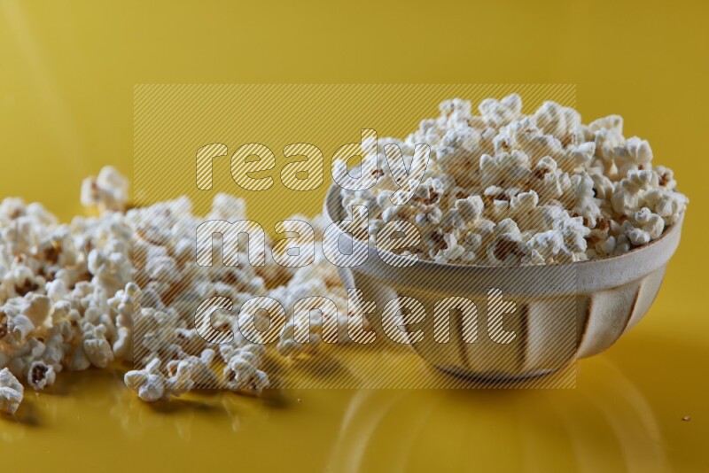 A white pottery bowl full of popcorn with popcorn beside it on a yellow background in different angles