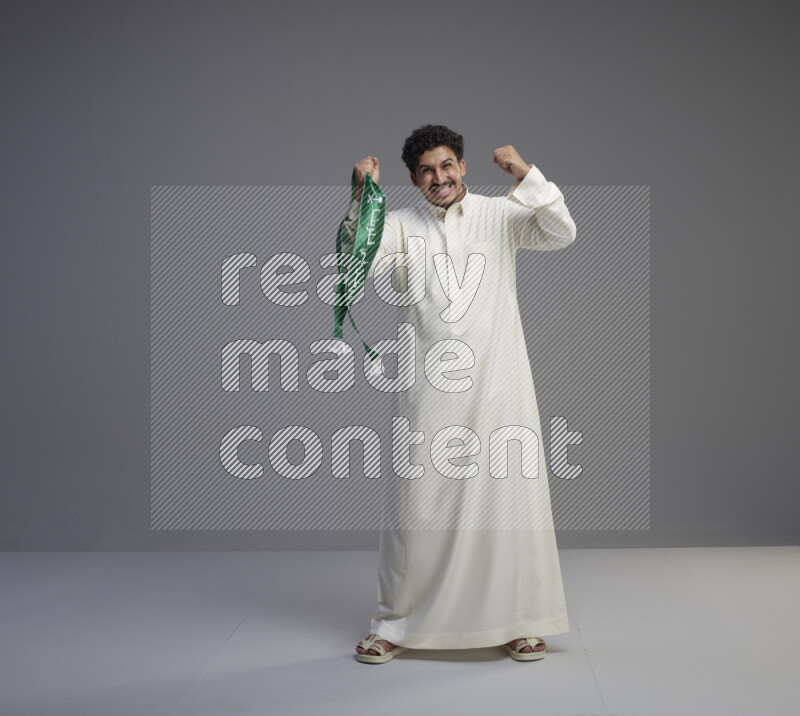 A Saudi man standing wearing thob holding Saudi flag scarf on gray background