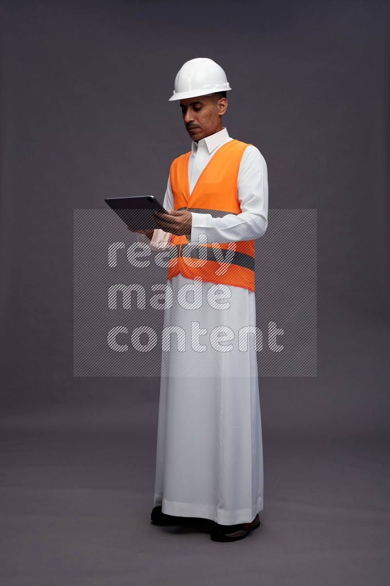 Saudi man wearing thob with engineer vest standing working on tablet on gray background