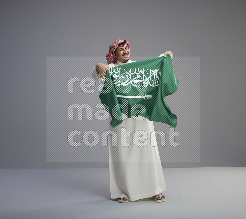 A saudi man standing wearing thob and red shomag holding big saudi flag on gray background