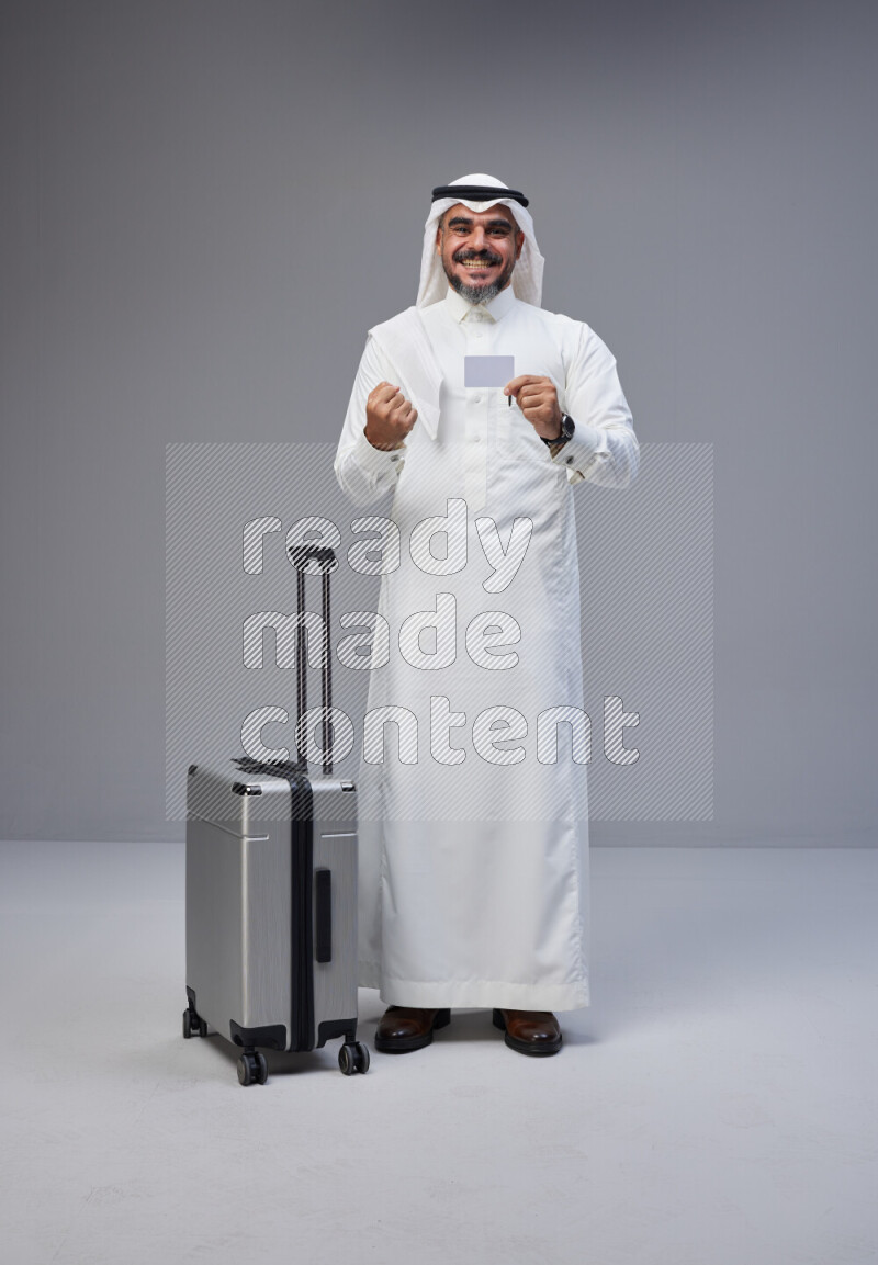 Saudi man wearing Thob and white Shomag standing holding Travel bag and ATM card on Gray background