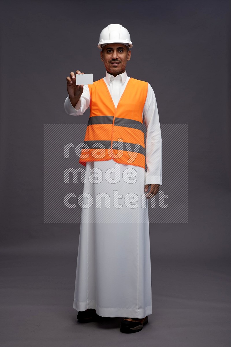 Saudi man wearing thob with engineer vest standing holding ATM card on gray background