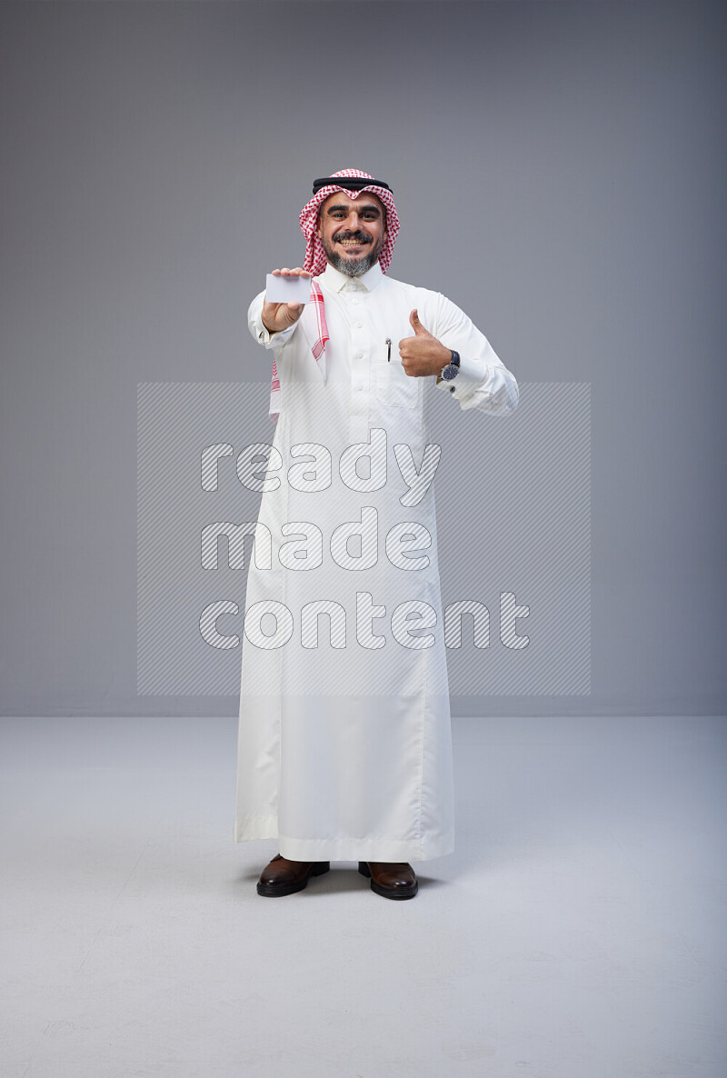 Saudi man Wearing Thob and red Shomag standing holding ATM card on Gray background