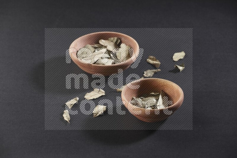2 wooden bowls full of dried bay leaves with more leaves spread on black flooring