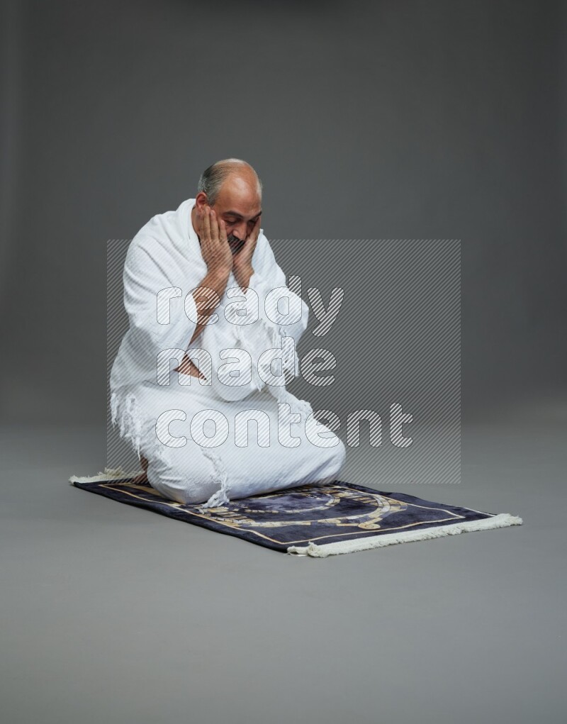 A man wearing Ehram sitting on prayer mat dua'a on gray background